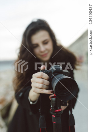 Female photographer adjusting professional camera on tripod outdoors by the sea, focusing the lens while preparing for a creative photo shoot on a rocky coastline. Female photographer adjusting professional camera on tripod outdoors by the sea, focusing the lens while preparing for a creative photo shoot on a rocky coastline. 134290427