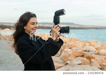 Young female photographer taking pictures outdoors by the sea, smiling while holding a professional camera with flash, capturing moments on a rocky coastline at sunset. 134290434