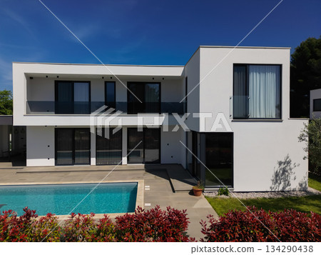 Modern luxury villa with swimming pool and clean minimalist architecture under a blue sky. Contemporary residential exterior featuring large windows, terrace, and elegant outdoor living space Modern luxury villa with swimming pool and clean minimalist architecture under a blue sky. Contemporary residential exterior featuring large windows, terrace, and elegant outdoor living space 134290438