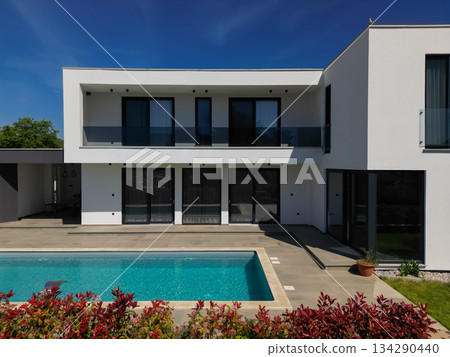 Modern luxury villa with swimming pool and clean minimalist architecture under a blue sky. Contemporary residential exterior featuring large windows, terrace, and elegant outdoor living space 134290440