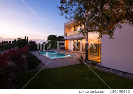Modern luxury villa with a luminous swimming pool at twilight. Contemporary architecture, sleek design, and inviting interior lights create a serene and elegant evening ambiance, ideal for upscale 134290480