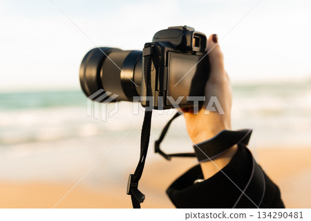 Close-up of woman holding a professional camera on the beach during golden hour, focusing on the lens and capturing outdoor photography moments. 134290481