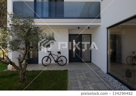 Modern residential entrance with minimalist architecture, front door, bicycle, and landscaped garden. Contemporary home exterior showcasing clean design, everyday lifestyle, and outdoor living 134290482