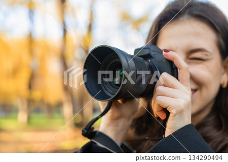 Young woman reviewing photos on her digital camera while standing in a sunny park. Warm autumn light, creative outdoor photography moment, lifestyle scene. 134290494