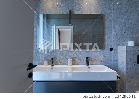 Modern, luxurious bathroom featuring a double vanity, sleek grey marble tiles, a walk-in shower, and a minimalist toilet. A mirror reflects a glimpse of the adjoining stylish bedroom 134290511