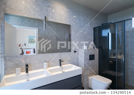 Modern, luxurious bathroom featuring a double vanity, sleek grey marble tiles, a walk-in shower, and a minimalist toilet. A mirror reflects a glimpse of the adjoining stylish bedroom 134290514