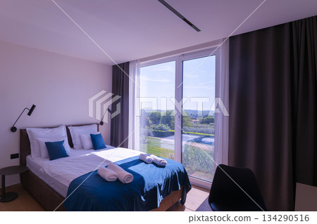 Modern bedroom with a comfortable bed, stylish blue accents, and a large window offering a serene view of lush greenery and distant horizons, perfect for relaxation and peaceful escapes 134290516