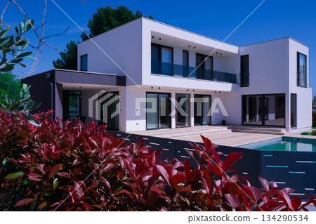 Modern luxury villa with swimming pool and clean minimalist architecture under a blue sky. Contemporary residential exterior featuring large windows, terrace, and elegant outdoor living space Modern luxury villa with swimming pool and clean minimalist architecture under a blue sky. Contemporary residential exterior featuring large windows, terrace, and elegant outdoor living space 134290534
