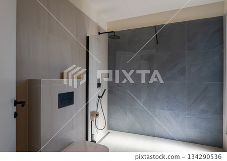 Modern, luxurious bathroom featuring a double vanity, sleek grey marble tiles, a walk-in shower, and a minimalist toilet. A mirror reflects a glimpse of the adjoining stylish bedroom Modern, luxurious bathroom featuring a double vanity, sleek grey marble tiles, a walk-in shower, and a minimalist toilet. A mirror reflects a glimpse of the adjoining stylish bedroom 134290536
