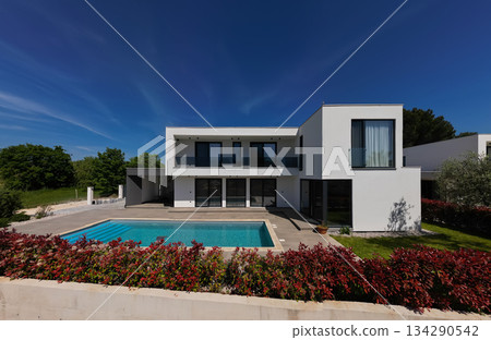 Modern luxury villa with swimming pool and clean minimalist architecture under a blue sky. Contemporary residential exterior featuring large windows, terrace, and elegant outdoor living space 134290542