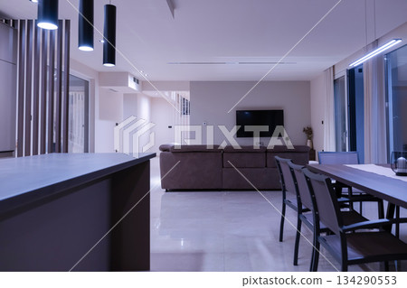 Interior of a modern luxury living room at night featuring a large L-shaped sofa, wall-mounted TV, and floor-to-ceiling windows overlooking an illuminated blue swimming pool. 134290553
