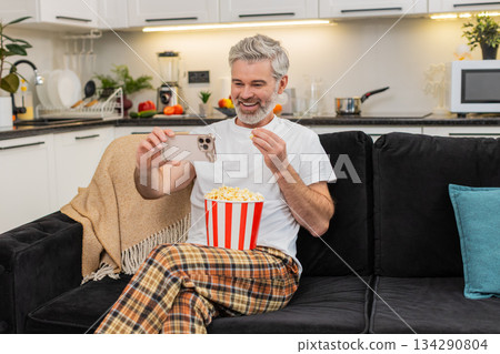 Middle-aged man switches shows on smartphone laughs eats popcorn watches online video relaxes fully 134290804