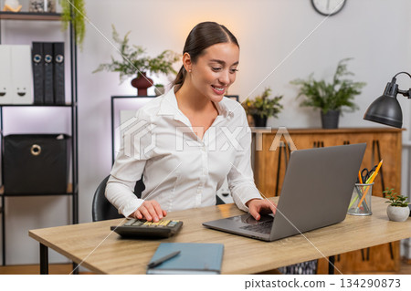 Young businesswoman manages budget in laptop spreadsheet clicks physical calculator and checks sums Young businesswoman manages budget in laptop spreadsheet clicks physical calculator and checks sums 134290873