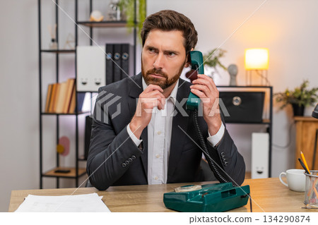 Middle-aged businessman lifts retro wired telephone and makes client call listening closely calmly Middle-aged businessman lifts retro wired telephone and makes client call listening closely calmly 134290874