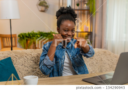 African American young woman flirts and shows call me hand gesture at camera sits at table at home African American young woman flirts and shows call me hand gesture at camera sits at table at home 134290943
