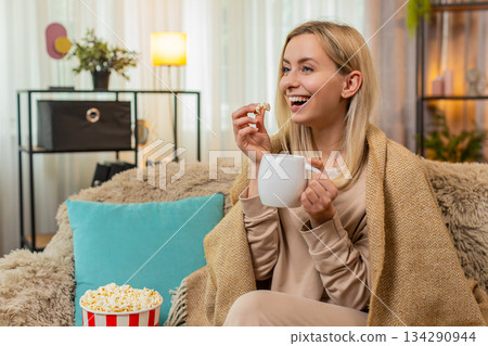 Adult woman wrapped in blanket sitting on sofa with cup of tea watching series thoughtfully at home Adult woman wrapped in blanket sitting on sofa with cup of tea watching series thoughtfully at home 134290944