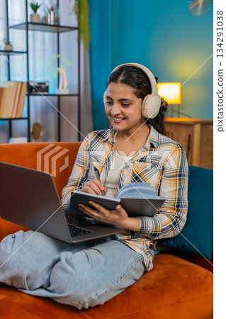 Indian woman listening music on laptop with headphones writing notes working freelance concentrated Indian woman listening music on laptop with headphones writing notes working freelance concentrated 134291038