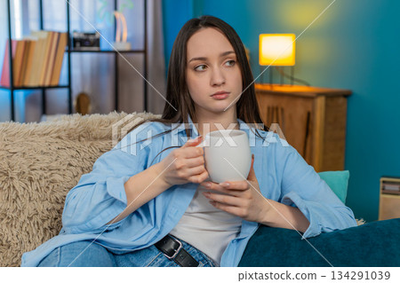 Sad young woman sitting on sofa drinking tea feeling lonely thoughtful mood melancholic atmosphere Sad young woman sitting on sofa drinking tea feeling lonely thoughtful mood melancholic atmosphere 134291039