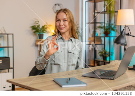 Happy Caucasian woman looking approvingly at camera showing OK gesture sitting at home office table Happy Caucasian woman looking approvingly at camera showing OK gesture sitting at home office table 134291138