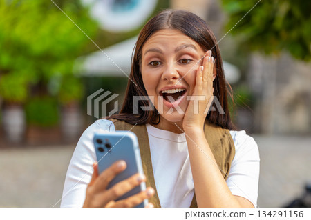 Excited European young woman using smartphone typing browsing celebrating win on urban city street 134291156