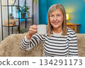 Happy mature woman raising coffee cup, smiling in greeting or gratitude cheers gesture on home sofa 134291173