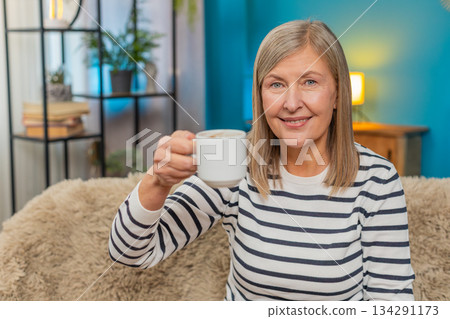 Happy mature woman raising coffee cup, smiling in greeting or gratitude cheers gesture on home sofa 134291173