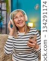 Mature woman listening to music on smartphone with wireless headphones dancing resting on home sofa 134291174