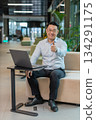 Happy young businessman working on laptop looking at camera showing thumbs up in modern office lobby 134291175