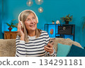 Mature woman listening to music on smartphone with wireless headphones dancing resting on home sofa 134291181