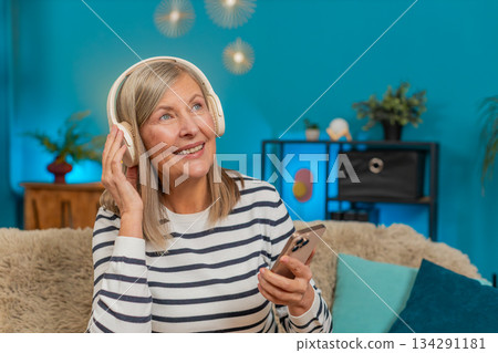 Mature woman listening to music on smartphone with wireless headphones dancing resting on home sofa 134291181