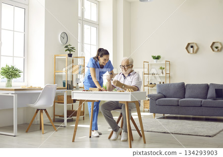 Nurse in retirement home helping senior man who is sitting at desk and doing puzzles Nurse in retirement home helping senior man who is sitting at desk and doing puzzles 134293903