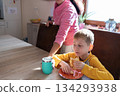 Young boy eating breakfast at the kitchen table while an adult places a drink nearby, morning daily routine and care. 134293938