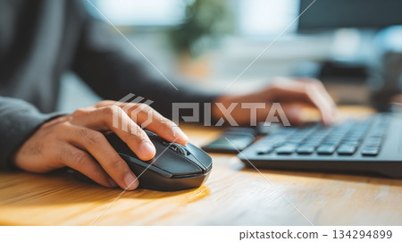 Hands operate a computer, using a mouse on a wooden surface and typing on a keyboard. Focus on efficiency and tech-driven work. Modern workplace, comfortable atmosphere. 134294899