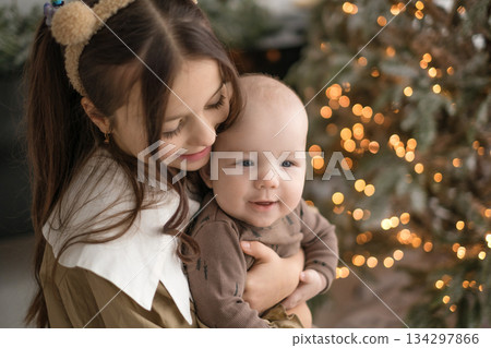 A girl holds her little brother in her arms against the background of a Christmas tree and the side lights of a garland. Waiting for New Year's miracles 134297866