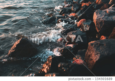 Water moves between rocks at the shoreline as waves crash softly while the sun sets in the background, creating a natural scene 134301689