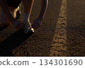 A person kneels to tie shoelaces on a quiet road as the sun sets in the background. The scene depicts an evening routine 134301690