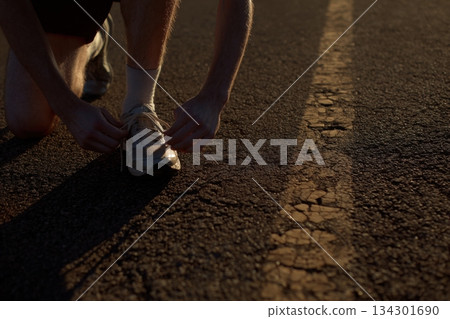 A person kneels to tie shoelaces on a quiet road as the sun sets in the background. The scene depicts an evening routine A person kneels to tie shoelaces on a quiet road as the sun sets in the background. The scene depicts an evening routine 134301690