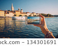 A person holds a phone to capture a city view by the water. The scene shows buildings and a boat under clear skies 134301691