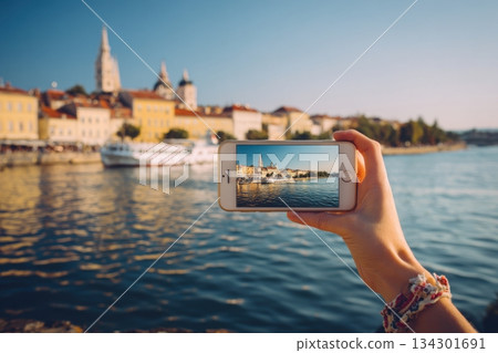 A person holds a phone to capture a city view by the water. The scene shows buildings and a boat under clear skies A person holds a phone to capture a city view by the water. The scene shows buildings and a boat under clear skies 134301691