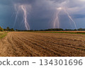Lightning illuminates the dark clouds above a field during a thunderstorm while rain begins to fall on the dry earth 134301696