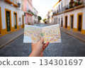 A person holds a colorful map in a quiet street of a historic town. The sun shines on the cobblestone road and nearby buildings 134301697