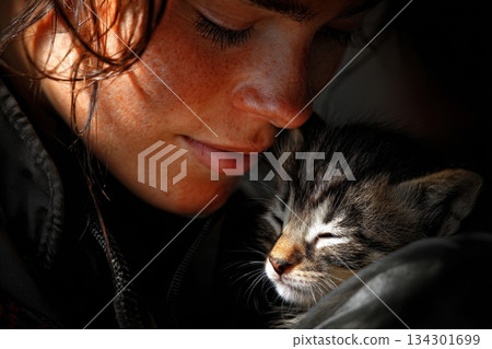 A person smiles while holding a small kitten close. The kitten appears relaxed and content in this moment A person smiles while holding a small kitten close. The kitten appears relaxed and content in this moment 134301699