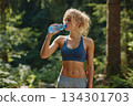 Woman stands in a forest, drinking water from a bottle while taking a break from her workout in nature 134301703