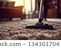 A person is cleaning the carpet with a vacuum cleaner in their living room while sunlight enters through the window 134301704