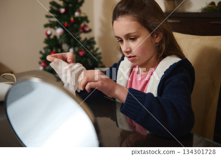 Young Girl Cleaning Up Tissue at Table With Christmas Tree in Background, Holiday Home Setting Young Girl Cleaning Up Tissue at Table With Christmas Tree in Background, Holiday Home Setting 134301728