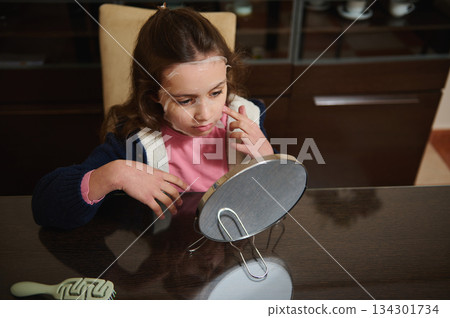 Young Girl With Facial Mask Looking Into Mirror At Kitchen Table For Self Care 134301734