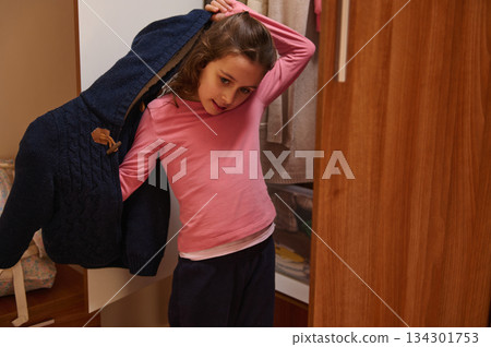 Little Girl Puts On A Cozy Jacket In A Bedroom Closet, Playfully While Getting Ready For The Day 134301753