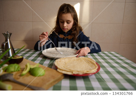 Young Girl Enjoying Pancakes At Home Kitchen Table With Avocado And Checkered Cloth Young Girl Enjoying Pancakes At Home Kitchen Table With Avocado And Checkered Cloth 134301766