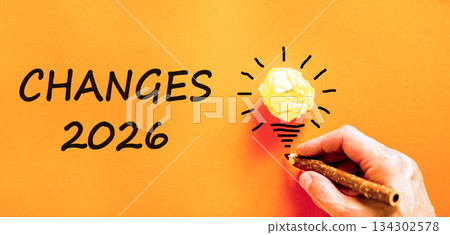 Changes 2026 symbol. Concept words Changes 2026 on beautiful orange paper. Beautiful orange paper background. Light bulb icon. Businessman's hand with pen. Business changes 2026 concept. Copy space 134302578