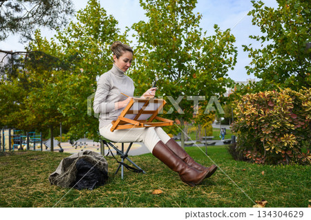 Woman painting on a portable easel with brushes in hand. Woman painting on a portable easel with brushes in hand. 134304629